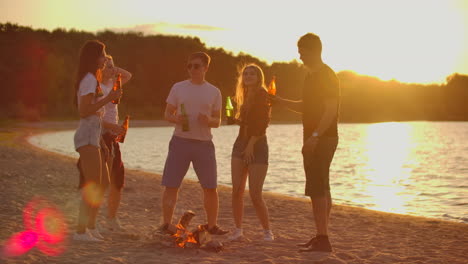 The-five-young-people-spend-time-at-sunset-on-the-beach-in-shorts-and-t-shirts-around-bonfire-with-beer.-They-are-dancing-and-enjoying-the-warm-summer-evening-near-the-river.